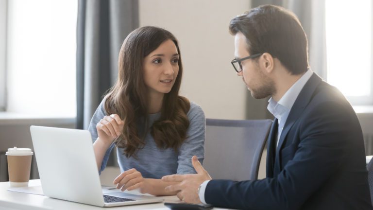 professional man and woman conversing while seated at a table with a laptop